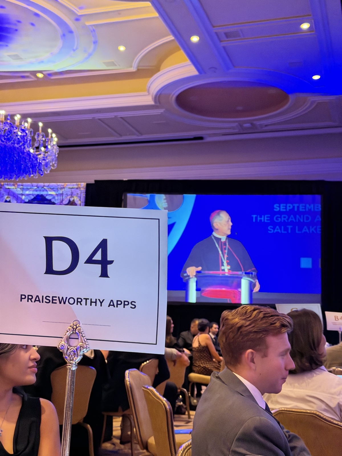 Praiseworthy Apps table card D4 at the 2026 Bishop's Dinner, with Bishop Oscar A. Solis speaking at the podium in the Grand America Hotel ballroom